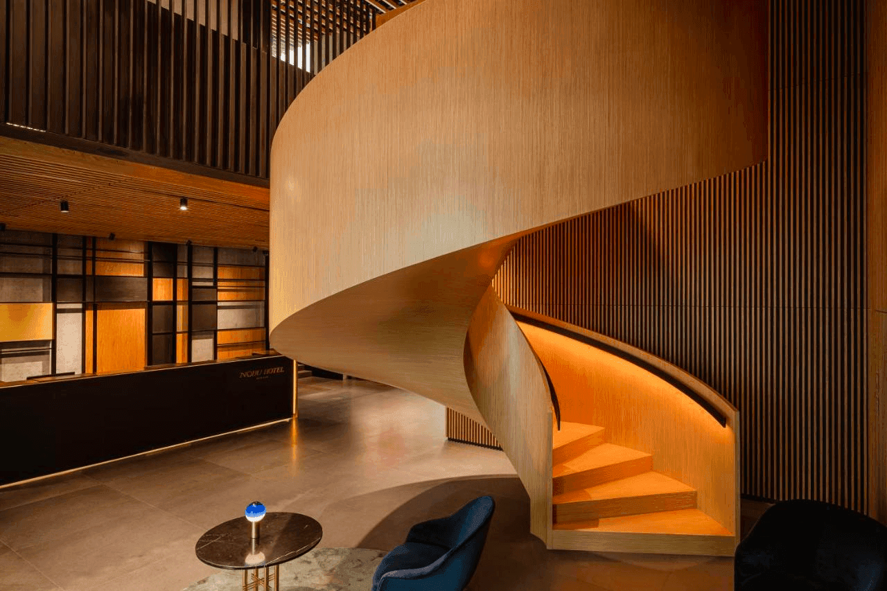 Interior of a hotel lobby with warm lighting, featuring a curved wooden staircase, vertical wooden slats on walls, a reception desk with the hotel's name, a round marble table with a blue glass ornament, and a navy blue chair.