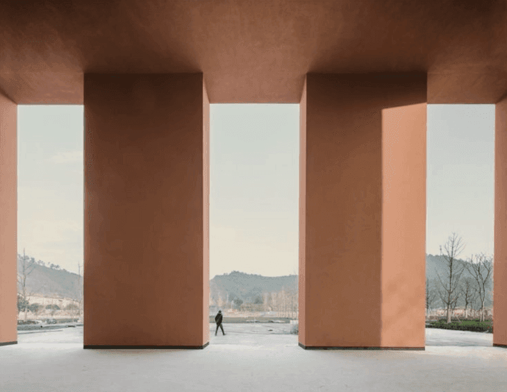 A modern architectural structure with terracotta-colored, rectangular columns, a person walking in the center, grassy landscape with trees in the background, overcast sky, and concrete flooring.