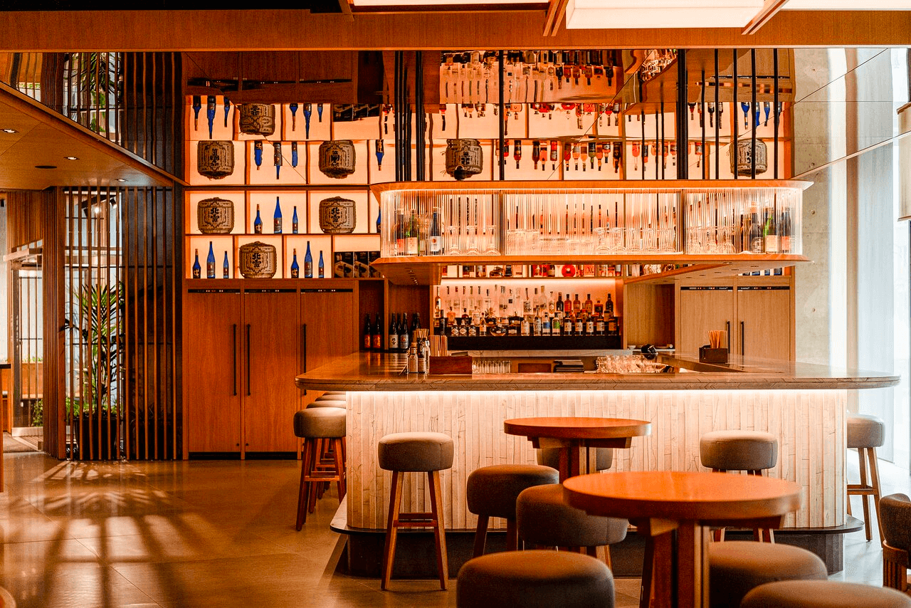 A cozy and stylish restaurant bar area with overhead glassware, wooden décor, shelves with various bottles, and framed artworks on the wall. There are round tables with stools, a wooden bar counter with lighting underneath, and a view of the outside through slatted wooden partitions.